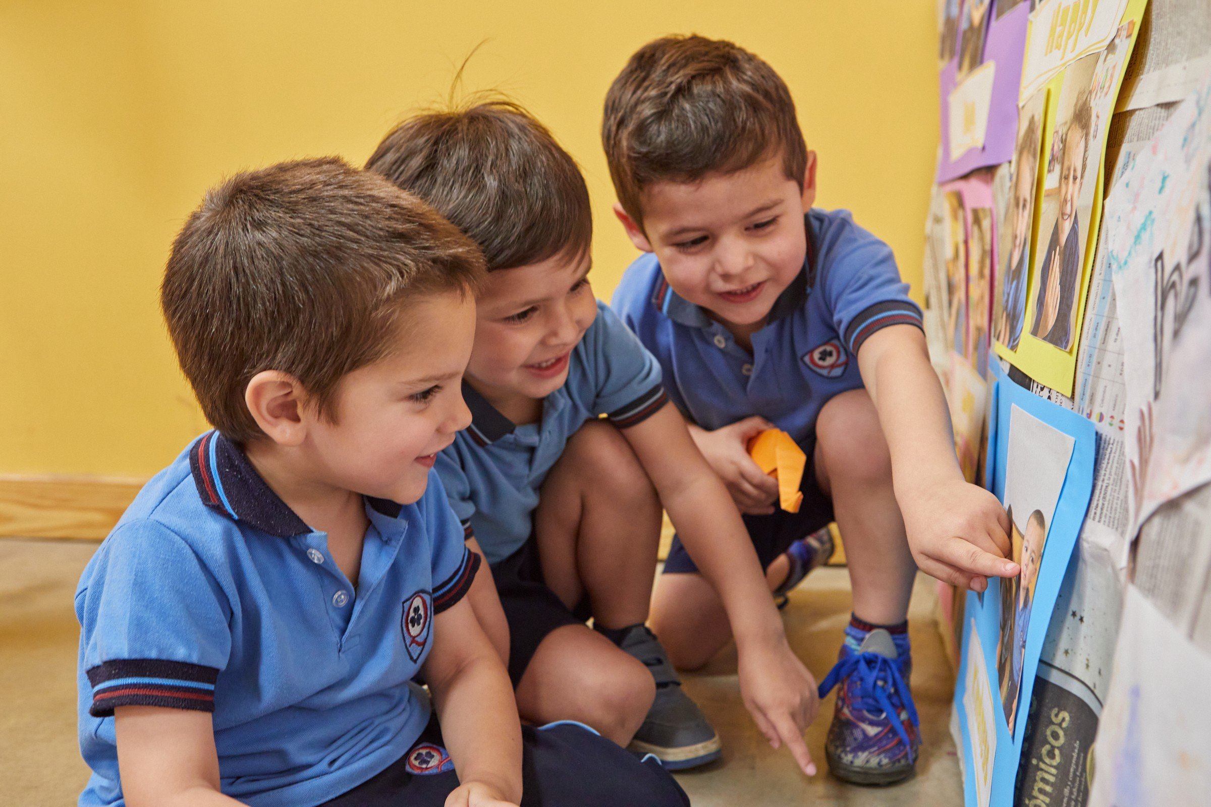 Tres alumnos de Nivel Inicial sonríen mientras uno señala una cartelera con fotos en la pared